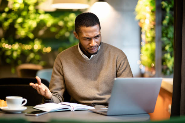 Surprised young black man looking at his computer Confused Guy. Afro manager looking at laptop, surprised by what he sees, spreading hands at modern cafe, free space confusion stock pictures, royalty-free photos & images