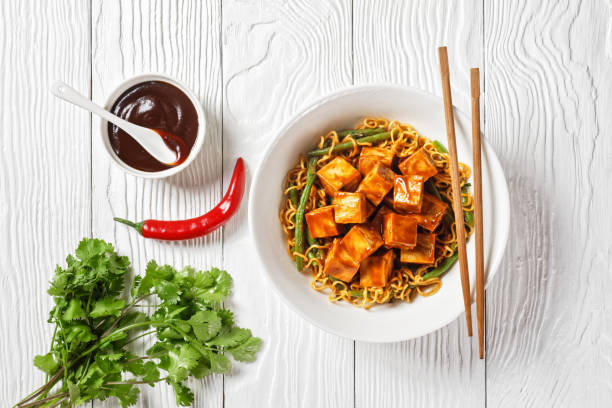 sweet and spicy asian noodle with hoisin baked crunchy tofu and  green bean  in a white bowl with chopsticks, horizontal view from above, flat lay stock photo