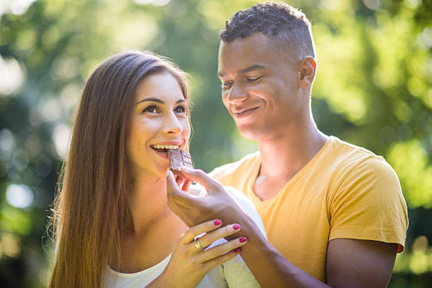 Sweet dating - chocolate and love Young man feeding woman with chocolate outdoors in park couple eating chocolate stock pictures, royalty-free photos & images