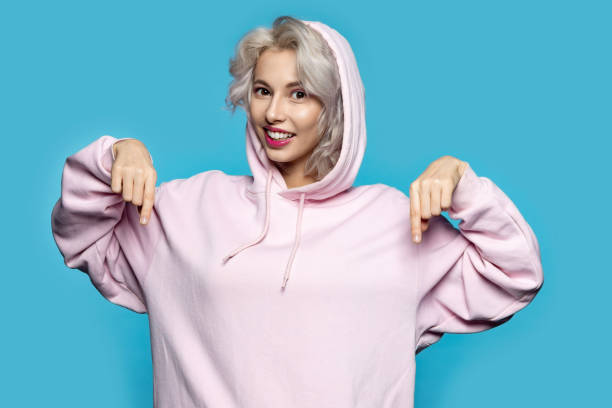 Sweet smiling female posing in studio Portrait of pretty happy lady showing on trendy light pink sweatshirt. Charming girl with short blonde hair dressed in stylish hoodie. Fashion concept. Isolated on blue hooded shirt stock pictures, royalty-free photos & images
