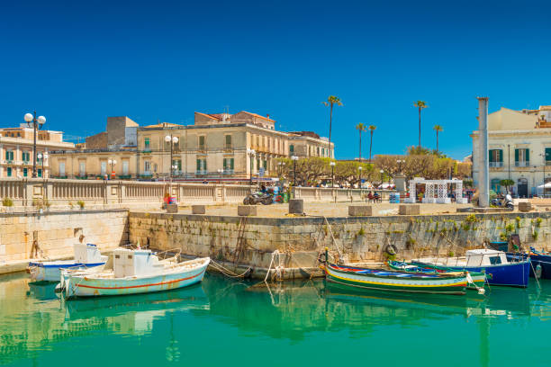 Syracuse, Italy: Cityscape of Syracuse and a small Island Ortygia, historical part of the city. Colorful boats lying on water near a stone pier stock photo