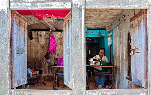 Bandipur, Nepal - April 23, 2022 : A tailor is sewing in his open workshop.  There are lots of traditional craft shops in this beautiful village where time seems to standstill. Many of them are owned by tailors and work with silk garments as  there is a silk factory in the town.
