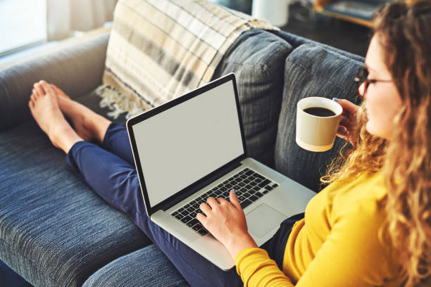 Foto seorang wanita muda menggunakan laptop dan minum kopi di sofa di rumah