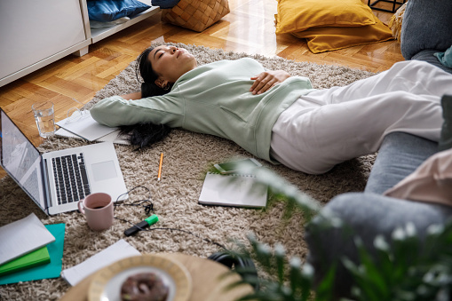 Three quarter length shot of female university student lying with hand behind head, on the living room floor, napping while taking a break from studying and doing homework.