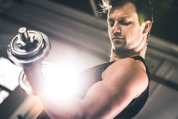 Taking care of his body Muscular young man doing exercises for biceps muscles with dumbbells on in a gym. Sun is shining from window in back. round dumbbells stock pictures, royalty-free photos & images