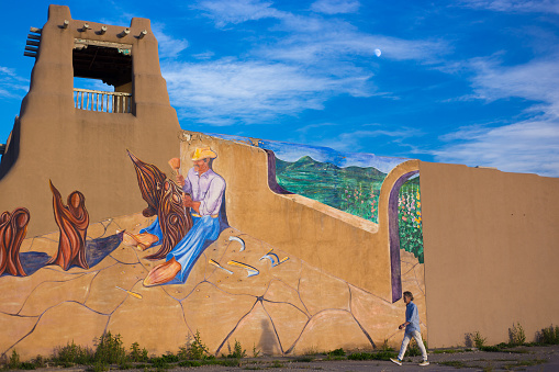 Taos, NM: A man walks past a mural on an adobe wall at dusk in downtown Taos. The mural shows a santero working on his wooden sculptures.