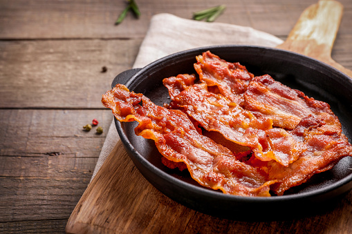 Hot fried crispy bacon slices in skillet on wooden background.