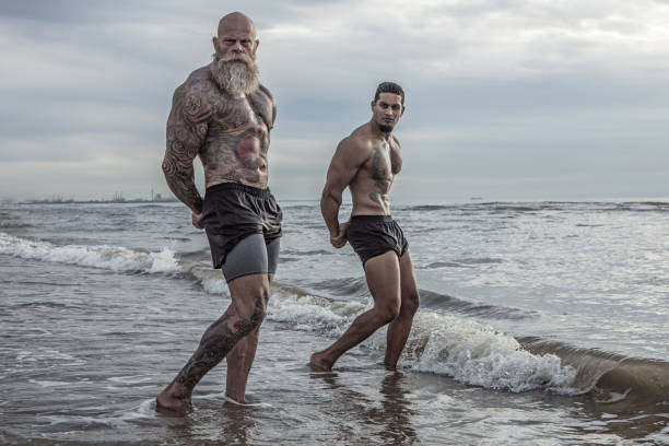 Tattooed Senior Man coaching young man During Workout Bearded Aggressive Redhead Senior Man coaching young man during outdoor workout on the beach male bodybuilders stock pictures, royalty-free photos & images