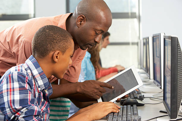 Teacher Helping Boy To Use Digital Tablet In Computer Class Teacher Helping Boy To Use Digital Tablet In Computer Class Having A Discussion computer training stock pictures, royalty-free photos & images