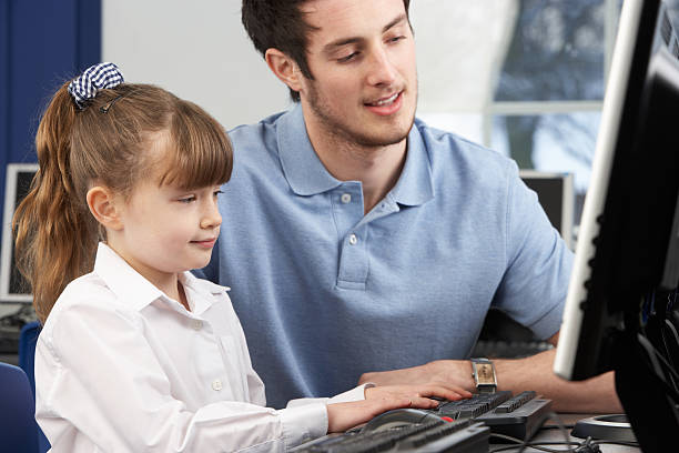 Teacher helping young girl in uniform how to use a computer stock photo