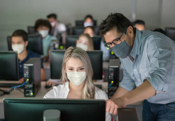 Teacher wearing a facemask and helping female student at an IT class Teacher wearing a facemask and helping female student at an IT class - education during the COVID-19 pandemic computer training stock pictures, royalty-free photos & images