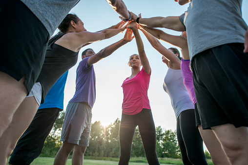 A multi-ethnic group of adults are taking an outdoor fitness class together at the park. They are standing together in a huddle and have their hands together in the middle.