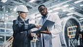 istock Team of Diverse Professional Heavy Industry Engineers Wearing Safety Uniform and Hard Hat Working on Laptop Computer. African American Technician and Female Worker Talking on a Meeting in a Factory. 1338031471