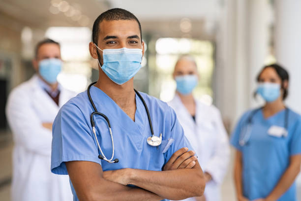 Team of doctors and nurses in hospital Confident multiethnic male nurse in front of his medical team looking at camera wearing face mask during covid-19 outbreak. Happy and proud indian young surgeon standing in front of his colleagues wearing surgical mask for prevention against coronavirus. Portrait of mixed race doctor with medical staff in background at hospital. medical occupation stock pictures, royalty-free photos & images