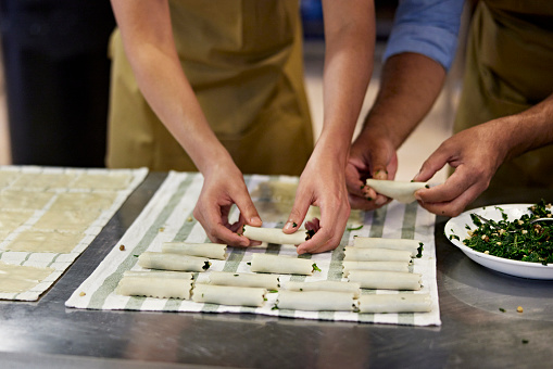 Men standing side by side at stainless steel workstation filling pasta squares with cooked spinach and rolling them up for baking.