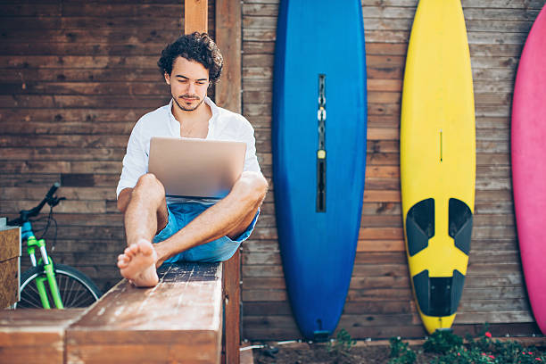 Technology and water sport Young man using laptop outdoors in the summer with surfing boards in the background. beach hut stock pictures, royalty-free photos & images
