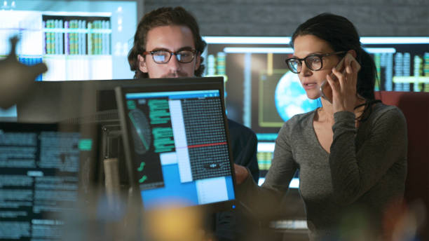 Technology displays man and woman Stock photo of a young man & woman working in an office surrounded by computer monitors displaying moving numerical & scientific data. online fraud stock pictures, royalty-free photos & images