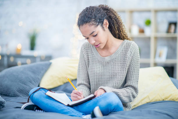 Teen girl journaling on her bed A pretty teenaged girl writes into a journal with interest while sitting cross legged on her bed. diary stock pictures, royalty-free photos & images