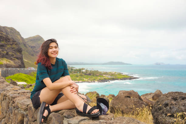 Teen girl sitting on rock wall overlooking Hawaiian ocean stock photo
