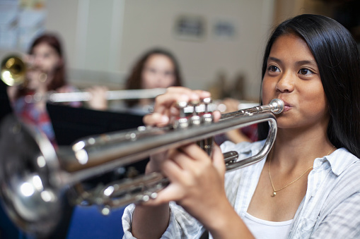 Confident Asian teenage girl blowing into trumpet in musical class. Students are learning music at high school. They are practicing woodwind instrument.