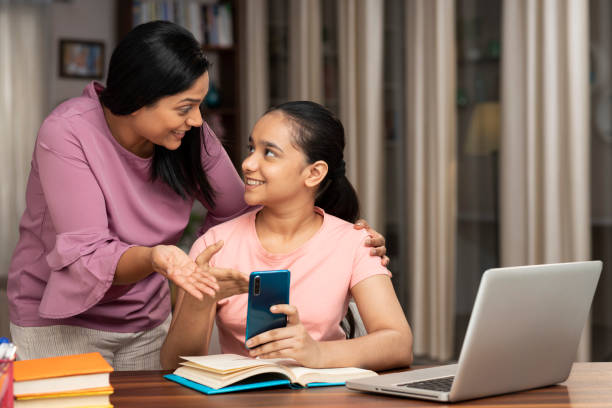 Teenage daughter showing mobile to her mom while studying at home stock photo