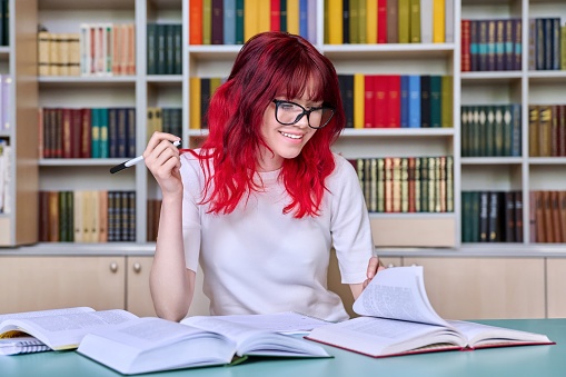 Teenage female student studying in library class, sitting at desk using books, writing in notebook. Fashion student with red dyed hair wearing glasses. College, study, education knowledge, adolescence
