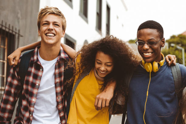 Teenage friends having fun outdoors Close up of three college friends standing in the street with arms around each other. Cheerful boys and a girl wearing college bags having fun walking outdoors. young adults hanging out stock pictures, royalty-free photos & images