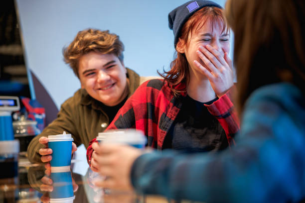 Teenage girl covering mouth while laughing, having coffee in paper cups with friends in cafeteria stock photo