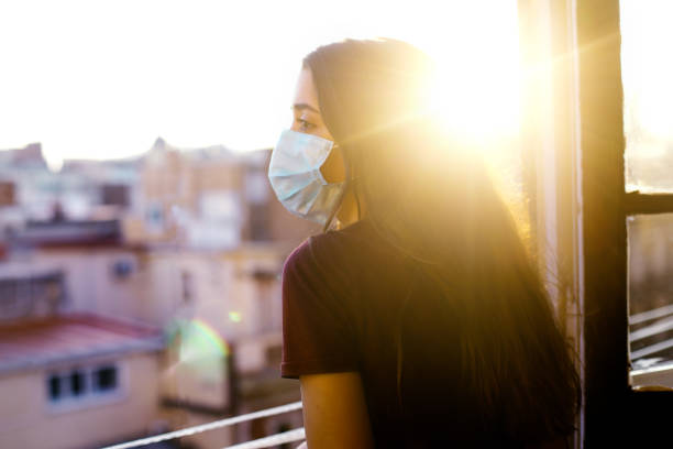 teenage girl in quarantine wearing protective mask looking out the window at sunset stock photo