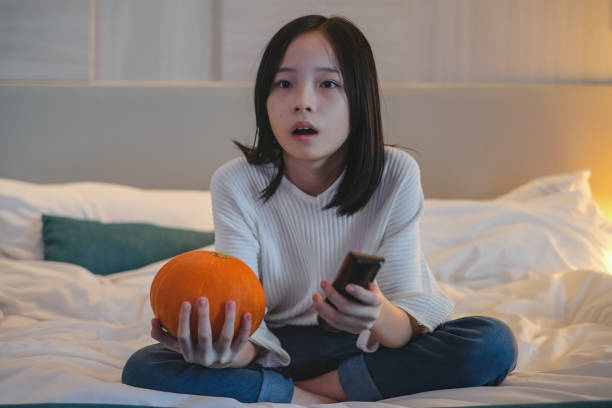 Teenage Girl Watching Horror Movie During Halloween Close-up shot of Asian teenage girl sitting on the bed looking terrified while watching a scary movie. asian kids watching tv stock pictures, royalty-free photos & images