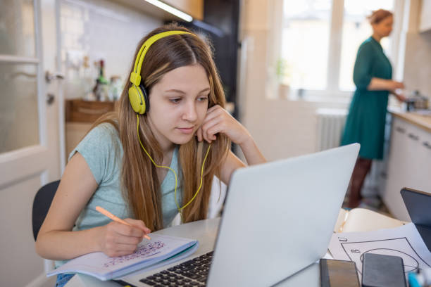 Teenage girl with headphones attending online class on laptop and taking notes stock photo