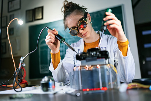 Teenage scientist girl wearing lab coat and steampunk goggles is working on her new invention - a mysterious power source that will solve humanity energy problems.
She is soldering the device.
Shot with Canon R5