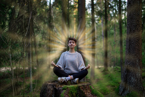 A teenager does yoga during the day in the woods. From it, bright sun rays go to the sides. Relaxation and tranquility. Eyes closed. Casual clothing. The concept of a healthy lifestyle.