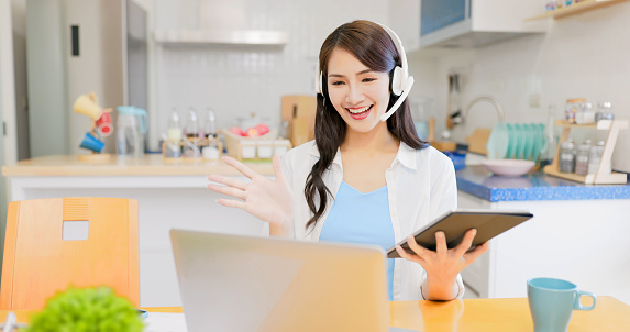 telework concept - asian brunette long hair businesswoman smiling with headset laptop computer and digital tablet in the kitchen at home
