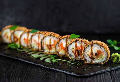 Japanese tempura hot sushi roll on Black slate stone plate on wooden background. sushi pieces with salmon, eel, cucumber, cream cheese, avocado wrapped in rice with crunchy seaweed on top with greens