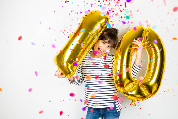 Ten birthday party girl with golden balloons and confetti Happy little girl holding two golden balloons making the 10 number while falling confetti on white background. 10th anniversary celebration party. 10 11 years stock pictures, royalty-free photos & images