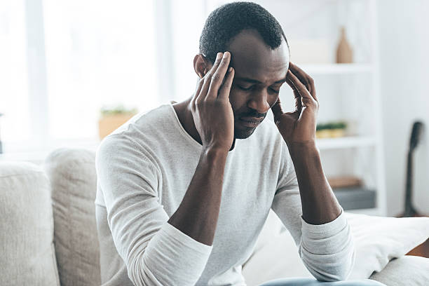 Terrible headache. Young African man touching head with hands and keeping eyes closed while sitting on the sofa at home headache stock pictures, royalty-free photos & images