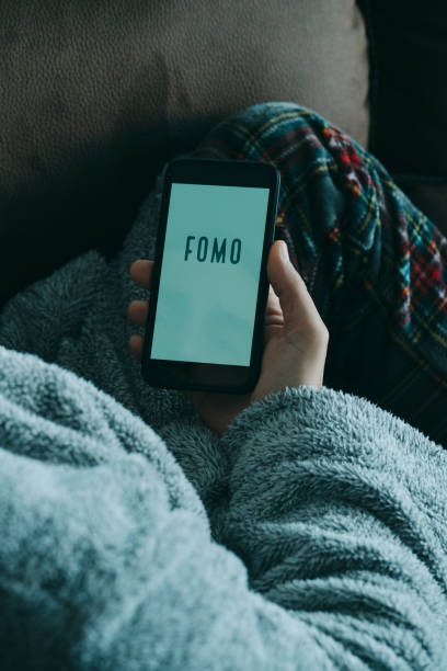 text fomo, for fear of missing out, in smartphone closeup a young caucasian man, wearing pajamas and house robe, lying in the sofa, watching his smartphone, that reads the text fomo, for fear of missing out, in its screen fomo stock pictures, royalty-free photos & images