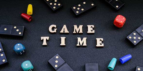 Text GAME TIME spelled out in wooden letter. Surrounded by dice, dominoes other game pieces on black background. Table games. Stay home activity for kids family.