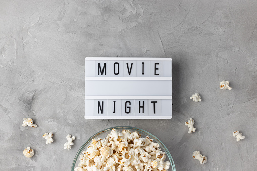 Text MOVIE NIGHT and popcorn in glass bowl on gray background. Cinema, movie theater, entertainment and snacks at home concept. Top view, copy space