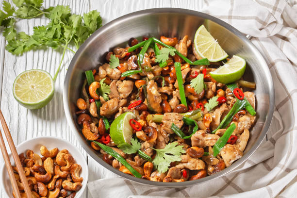 Thai cashew chicken stir fry in a pan on a wooden table with chopsticks and ingredients, horizontal view from above, close-up stock photo