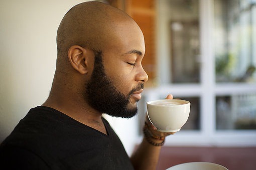 A 20-24-year-old African young male with a shaven bald head and black beard side view portrait enjoying a cup of coffee.  He enjoys the coffee and is smiling.  He is sitting on an outside porch of a restaurant or at home.  He looks happy and relaxed.
