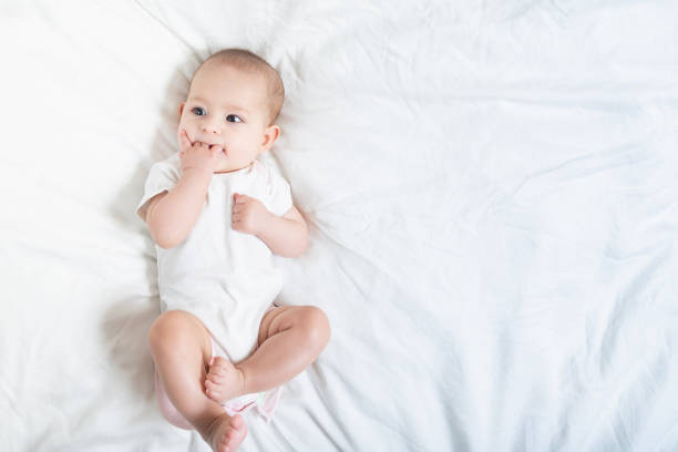 The baby teething Close up portrait of little baby girl in white lying on a white bed. stock photo
