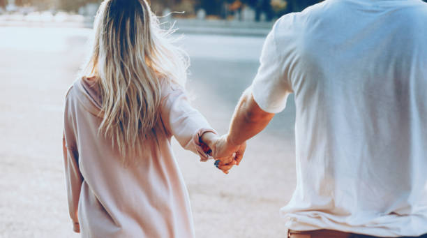 The back view of a young caucasian couple dressed in white walking hand in hand during sun is warming them The back view of a young caucasian couple dressed in white walking hand in hand during sun is warming them bonding stock pictures, royalty-free photos & images