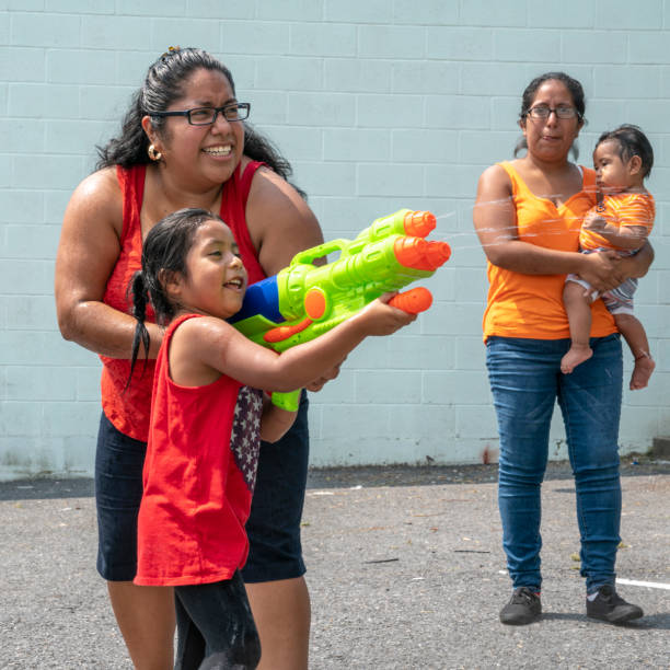 The big happy Latino, Mexican-American family - the mother, body-positive optimistic smiley woman, her sister, and kids, girls of different ages - playing outdoor with a water gun and having fun The big happy Latino, Mexican-American family - the mother, body-positive optimistic smiley woman, her sister, and kids, girls of different ages - playing outdoor with a water gun and having fun in the sunny hot summer day at the parking lot nearby his house in Pennsylvania, USA hot mexican girls stock pictures, royalty-free photos & images