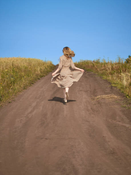 The blonde runs down a country road in a striped dress from the back stock photo