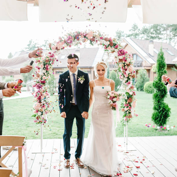 The bride and groom go through the arch, theirs sprinkled with leaves of roses and colored confetti. Wedding ceremony decorated with flowers and greenery of the outdoor in backyard banquet area. stock photo