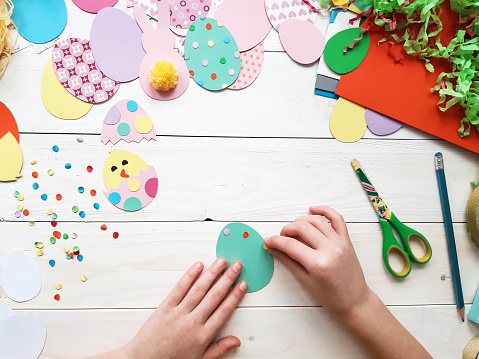 The child makes crafts with his own hands for the Easter. Colorful handmade from multi-colored paper. Scissors, cardboard, eggs, chicken, rabbit. Art creativity on a wooden table. Top view, copy space