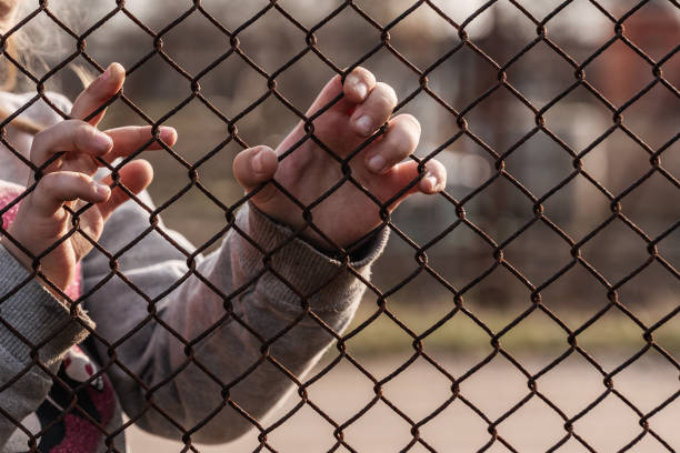 The child's hands are holding onto a metal mesh fence. Social problem of refugees and forced migrants stock photo