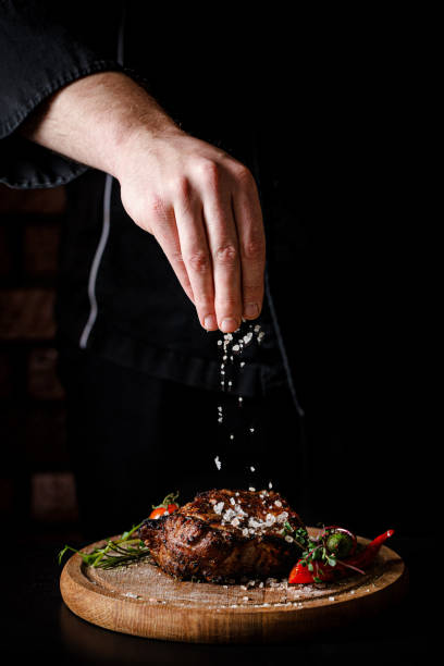 The concept of cooking meat. The chef cook salt on the cooked steak on a black background, a place under the logo for the restaurant menu. food background image, copy space text stock photo